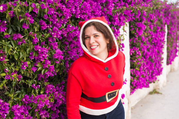 Christmas, holidays, humor and people concept - young happy woman smiling in christmas suit at summer near the purple flowers - Photo, Image