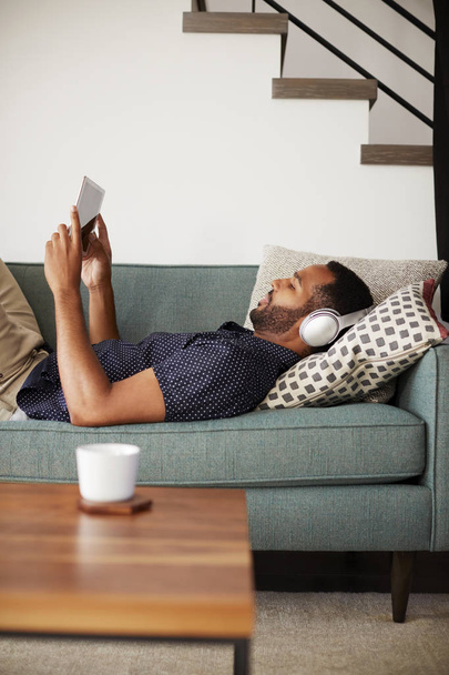 Man Lying On Sofa At Home Wearing Headphones And Watching Movie On Digital Tablet - 写真・画像