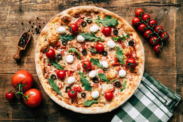 Italian pizza with tomatoes, mozzarella cheese, olives and arugula on old wooden background, top view - Fotó, kép