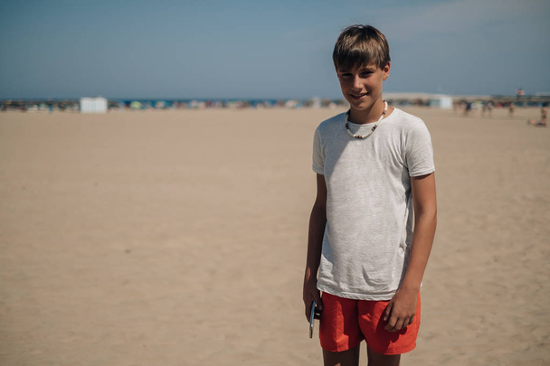 Chłopiec stoi na słonecznej plaży i uśmiechając się trzymając telefon komórkowy - Zdjęcie, obraz