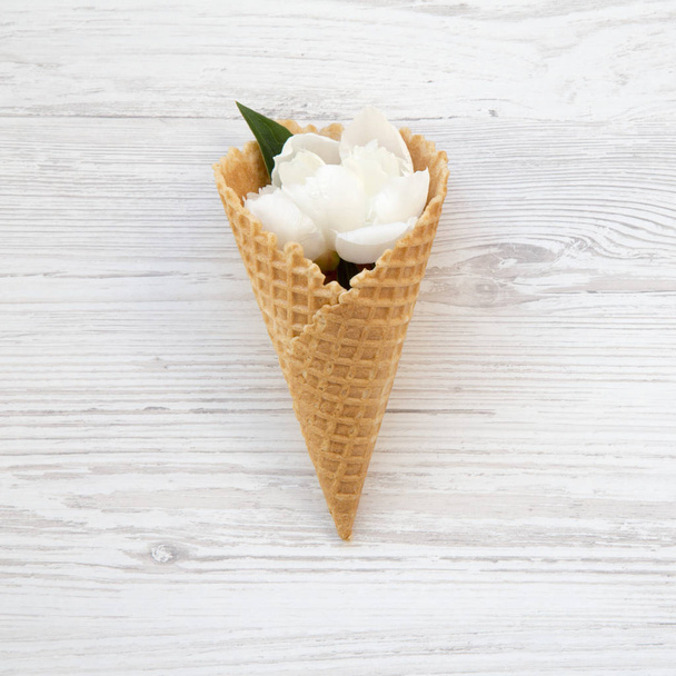 Sweet waffle cone with white peony flowers on a white wooden background, top view. - Фото, изображение