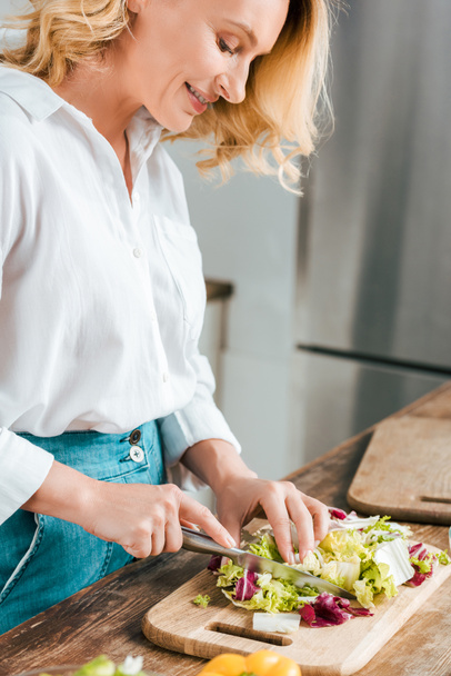 close-up shot of happy adult woman cutting lettuce for salad at kitchen - Фото, изображение