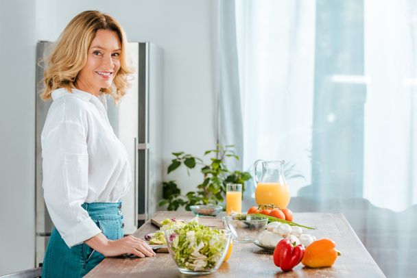 side view of happy adult woman looking at camera while making salad at kitchen - Fotó, kép