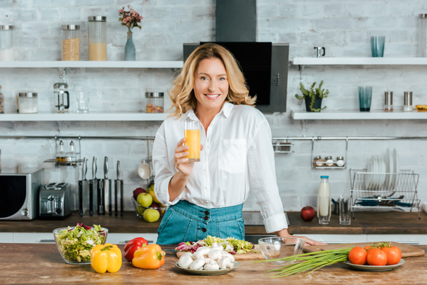 beautiful adult woman drinking orange juice while cooking at kitchen and looking at camera - Zdjęcie, obraz