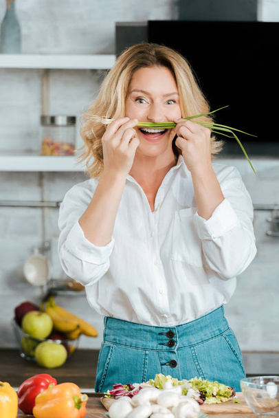 funny adult woman attaching leek like mustache at kitchen and looking at camera - Foto, Imagem