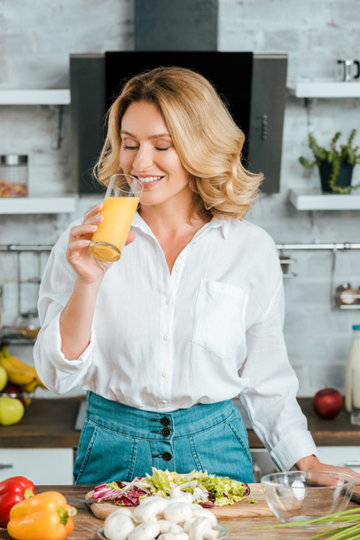 όμορφη ενήλικη γυναίκα πίνοντας χυμό πορτοκαλιού, ενώ το μαγείρεμα στην κουζίνα - Φωτογραφία, εικόνα