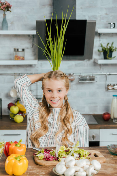 happy little child holding bunch of leek behind head while making salad at kitchen and looking at camera - Zdjęcie, obraz