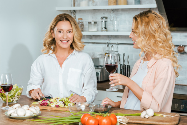 happy mature mother and adult daughter with glasses of red wine cooking together at kitchen - Фото, зображення