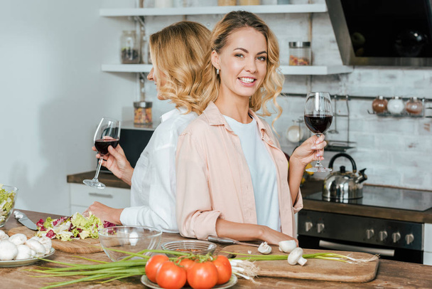 madre madura y sonriente hija adulta con copas de vino tinto de pie espalda con espalda en la cocina
 - Foto, Imagen