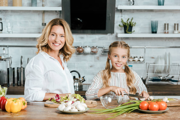 smiling adult mother and little daughter cooking together and looking at camera - Fotoğraf, Görsel