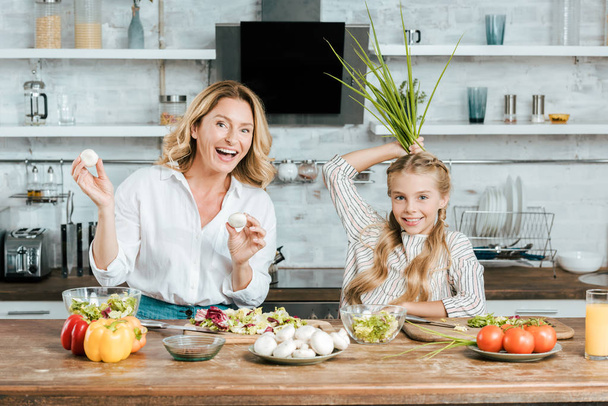 šťastný matka a dcera hrát s potravinami při vaření při pohledu na fotoaparát doma - Fotografie, Obrázek