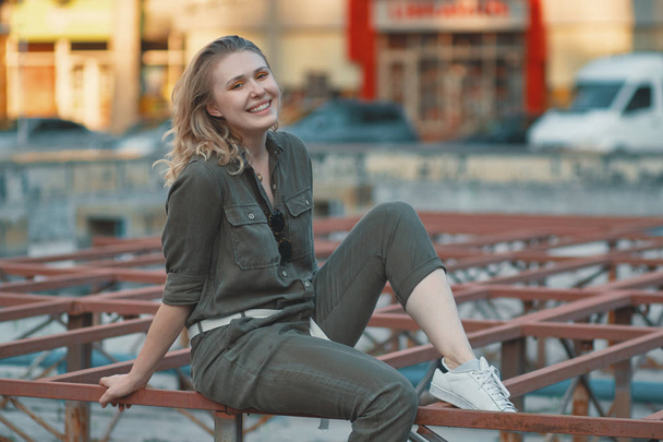 Porträt eines lässigen blonden Mädchens, das auf der Metallkonstruktion sitzt, mit einem Gebäude im Hintergrund. junge moderne Frau posiert in der Stadt, im Freien. Stadtleben - Foto, Bild