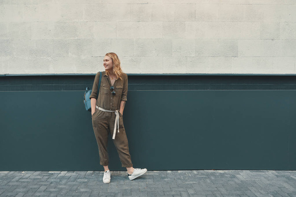 A hipster girl with long blonde hair, wearing a stylish romper and a backpack is looking aside while standing on a wall background on a street. Horizontal mock up. Empty space for text or design. - Fotoğraf, Görsel