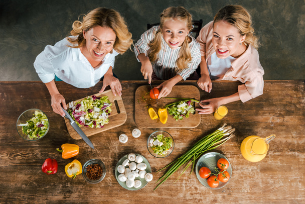 overhead view of child cutting vegetables for salad with mother and grandmother and looking at camera - Photo, Image