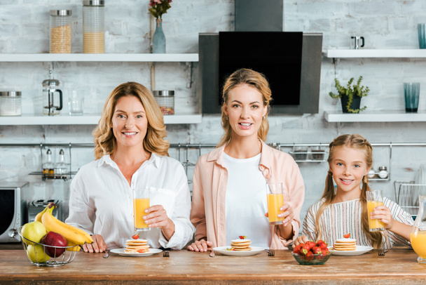 mooie drie generaties van vrouwen met pannenkoeken en sinaasappelsap bij het ontbijt thuis kijken naar camera - Foto, afbeelding