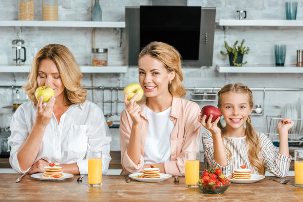 Gelukkig klein kind met moeder en grootmoeder met pannenkoeken en gezonde appels voor ontbijt thuis kijken naar camera - Foto, afbeelding