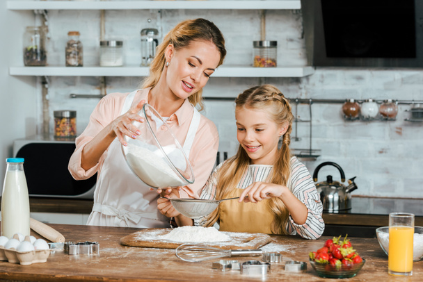 ευτυχισμένη μητέρα και κόρη ρίχνει το αλεύρι μέσα από κόσκινο για ζύμη στην κουζίνα - Φωτογραφία, εικόνα