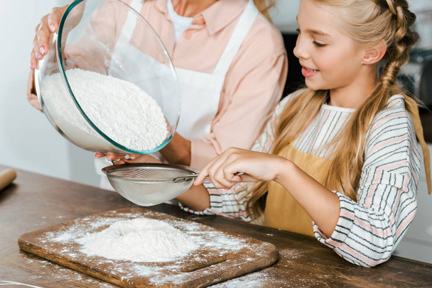 cropped shot of mother and daughter pouring flour through sieve for dough at kitchen - Foto, imagen