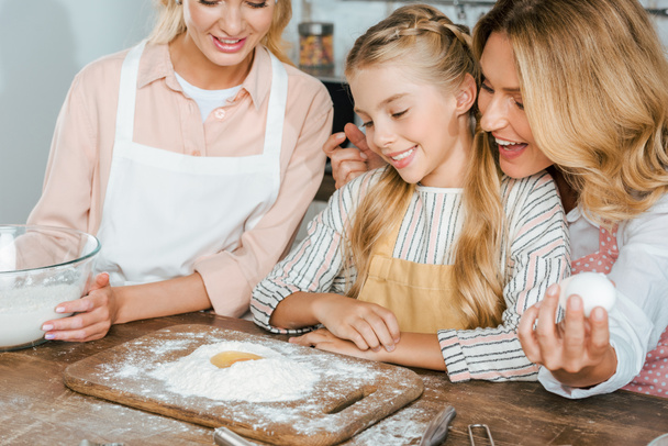 close-up shot of little child with mother and grandmother making dough with flour and egg together at home - Photo, Image