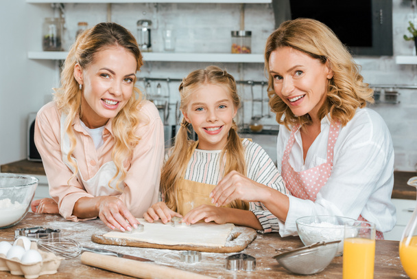 piękny szczęśliwy trzech pokoleń kobiet, wyrabiania ciasta razem w domu i patrząc na kamery - Zdjęcie, obraz