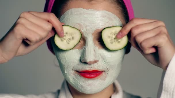 Close up of a girl with a face mask and a towel on her head that closes her eyes with cucumbers on a gray background. Face care concept. Beautiful girl with cosmetic mask on her face. - Imágenes, Vídeo