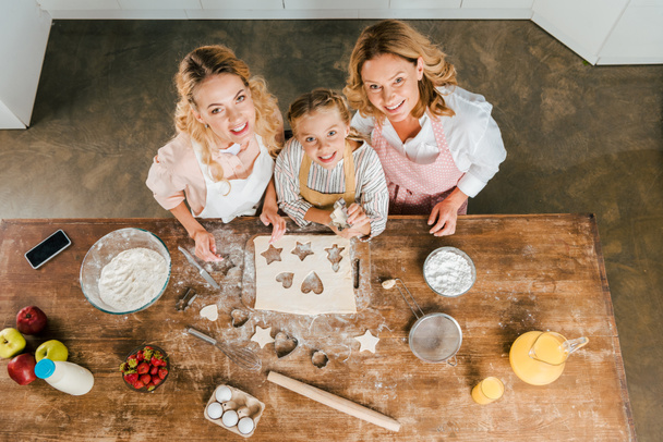top view of smiling child with mother and grandmother preparing christmas cookies at home and looking at camera - Photo, Image