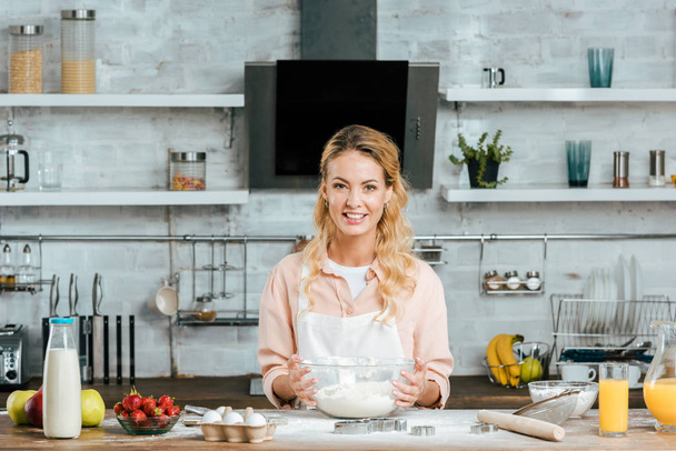 souriant jeune femme avec bol de pâte regardant caméra à la cuisine
 - Photo, image