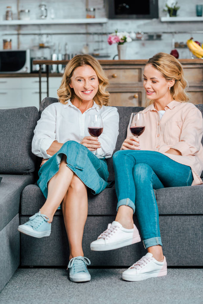 mature mother and adult daughter with glasses of red wine spending time together on couch at home - Photo, Image