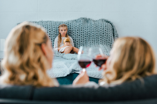sad lonely child sitting on bed with toy while her mother clinking glasses of wine with friend on foreground - Photo, Image