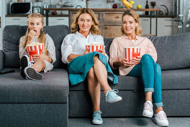 happy child with mother and grandmother watching movie on couch at home with buckets of popcorn - Photo, Image