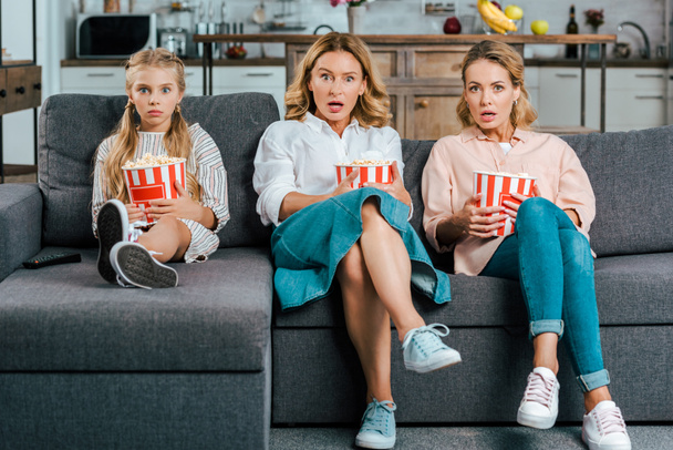 шокированные ребенок с матерью и бабушкой смотреть кино на диване дома с ведрами попкорна
 - Фото, изображение