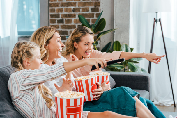 excited child with mother and grandmother watching movie on couch at home with buckets of popcorn and pointing at screen - Zdjęcie, obraz