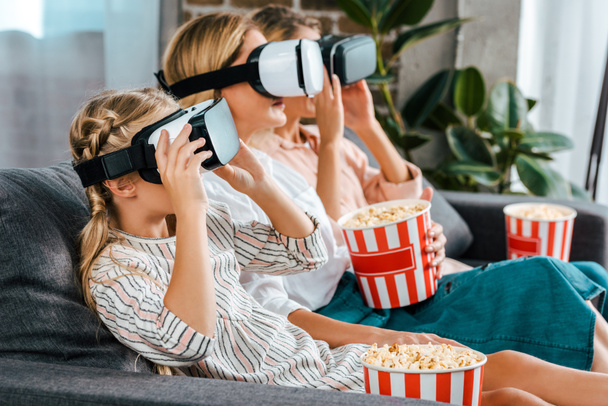 side view of child with mother and grandmother sitting on couch together and watching movie with virtual reality headsets at home - Фото, зображення