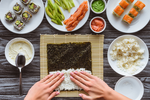 tiro cortado de pessoa que prepara sushi com arroz e nori, ingredientes gourmet na mesa
  - Foto, Imagem