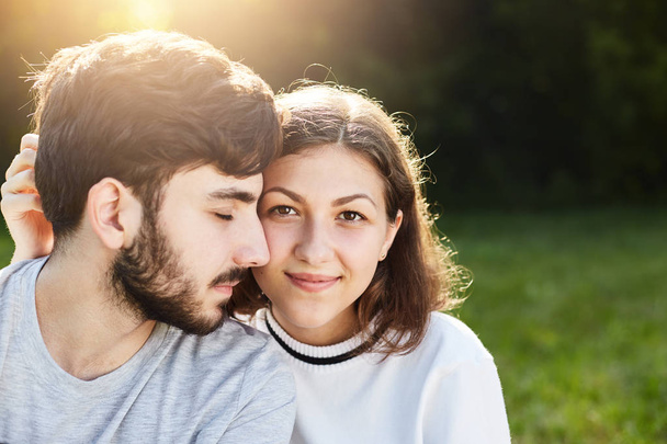 Candid shot of lovely couple admiring every minute spending together sitting at green grass of meadow. Carefree guy with dark hair and thick beard closing his eyes with pleasure to be near his lover - Foto, Imagen