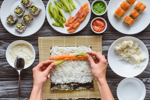 colpo ritagliato di persona che cucina delizioso rotolo di sushi con salmone, riso, cetriolo, avocado e nori
  - Foto, immagini