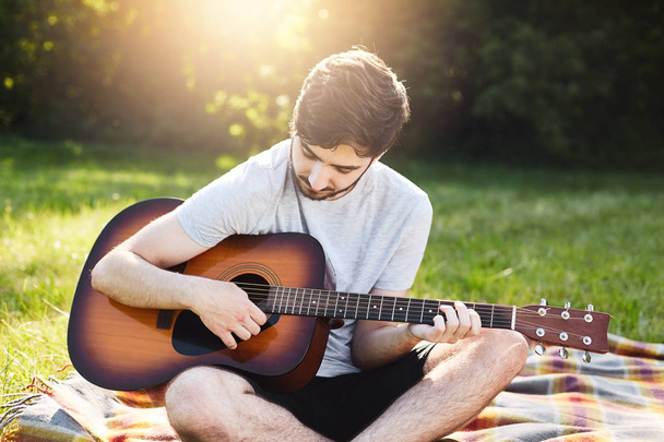 Carefree male with dark beard and trendy hairstyle learning to play guitar while sitting crossed legs at wrap. Young man playing musical instrument at green nature. Music, hobby and rest concept - Fotografie, Obrázek