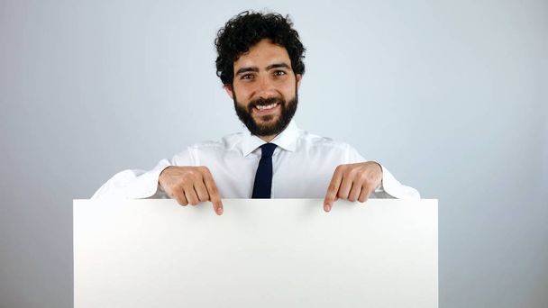 Bonito homem com uma expressão sorridente segurando um banner em branco e apontando um espaço de cópia para o seu texto ou publicidade, sobre um fundo cinza
 - Foto, Imagem