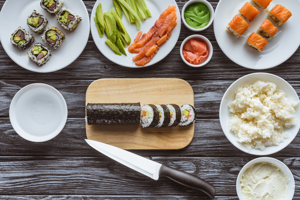 top view of delicious sliced sushi roll, knife and ingredients on wooden table  - Foto, afbeelding