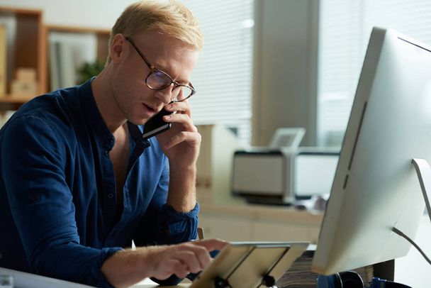 Joven hombre de negocios en gafas usando tableta y llamando por teléfono
 - Foto, imagen