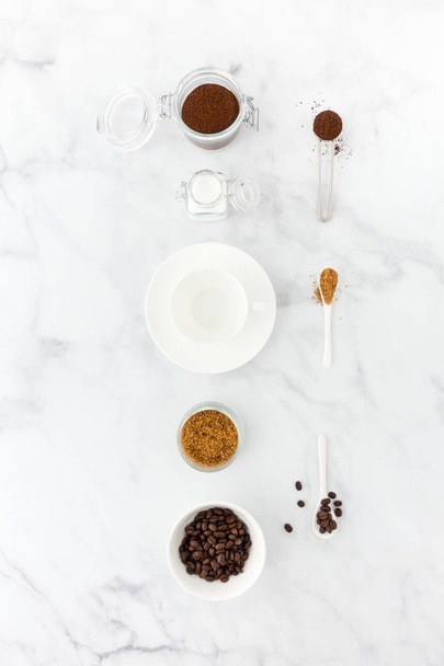 Granos de café, café molido, azúcar moreno y blanco, cuchara de medir café y una taza de café vacía sobre fondo de mármol blanco
. - Foto, Imagen