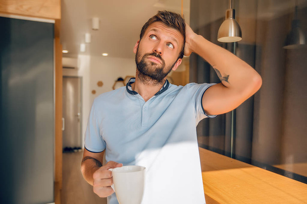 красивий бородатий чоловік посміхається тримає білу чашку кави, стоячи на кухні
 - Фото, зображення