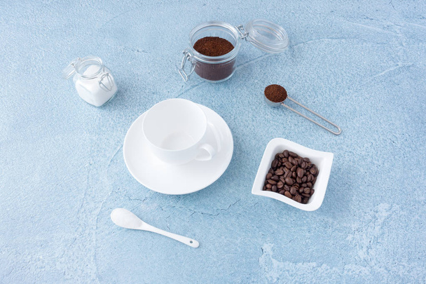Conceito de café com copo de café vazio, açúcar branco e café moído em frascos de vidro no fundo de concreto azul
. - Foto, Imagem