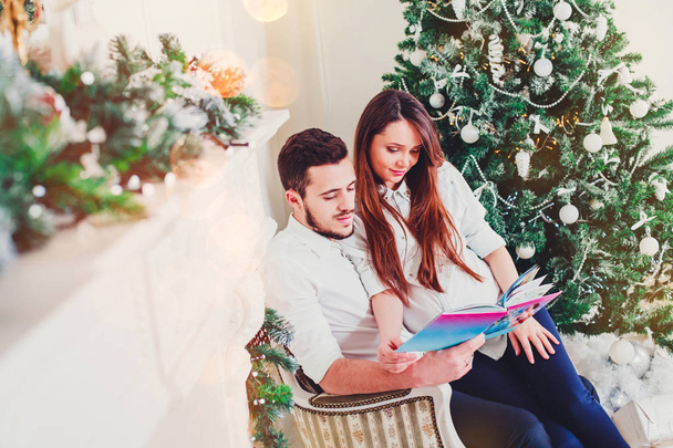 Boldog pár ül a nappaliban, amelyek a karácsonyi könyv, a díszített karácsonyi fa mögött a fény ad egy barátságos légkörben. Szilveszter és a karácsony téma - Fotó, kép