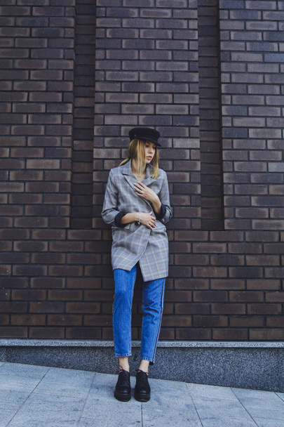 Young beautiful girl in a jacket, jeans and hat on a brick wall background - Фото, зображення