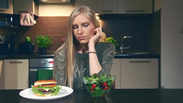 Jonge vrouw kiezen tussen hamburger en salade. Mooie jonge dame op zoek naar heerlijke hamburger en gezonde groente salade zittend aan tafel in de stijlvolle keuken. - Video