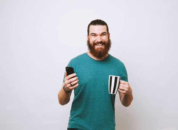 Happy smiling bearded man holding cup of coffee and smartphone and looking at the camera - Photo, image