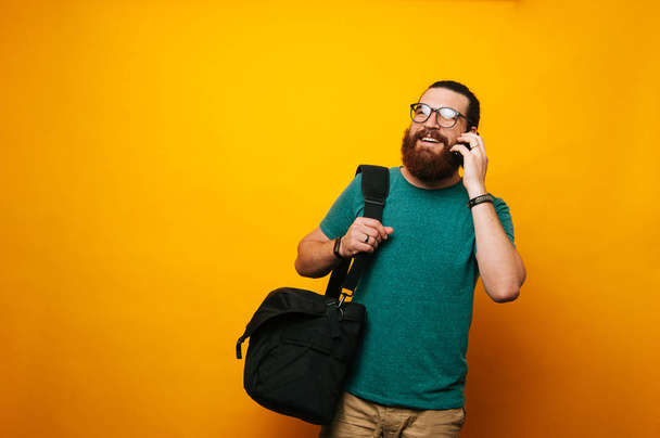 Веселий молодий бородатий чоловік розмовляє по телефону збирається працювати зі своєю сумкою
. - Фото, зображення