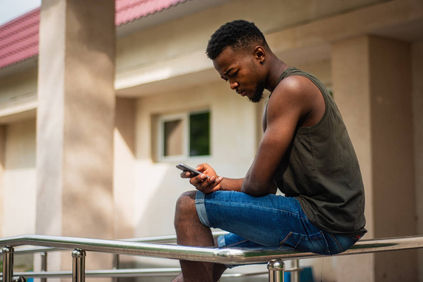 Concentrated urban hipster, a young man using a smartphone. African American teenager holding a mobile smartphone sitting on the railing - Foto, Imagem