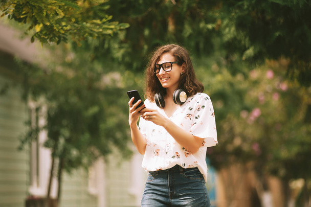 Photo of happy young woman walking outdoors in park. Looking aside listening music while chatting by phone. - Foto, afbeelding